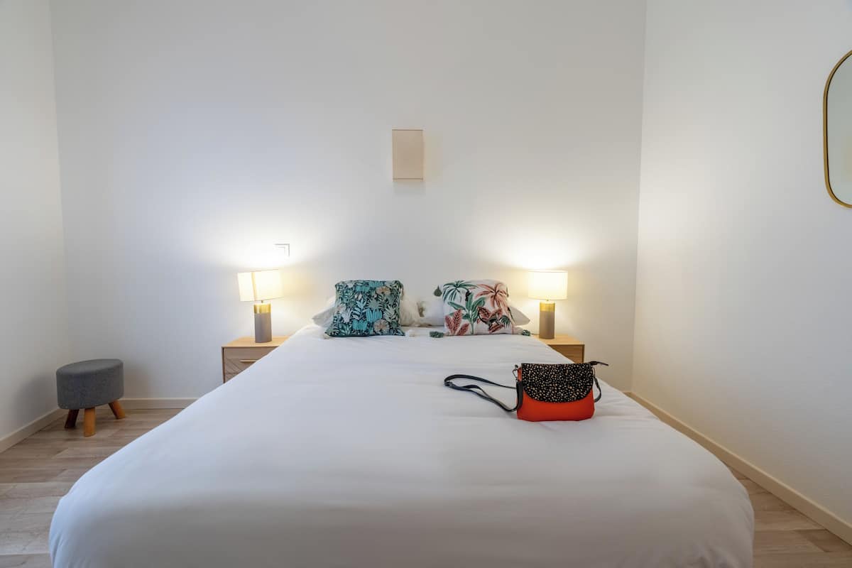 A neatly made bed with two pillows, a small stool, and a handbag on the bed. There are two bedside lamps and a small table with a lamp.