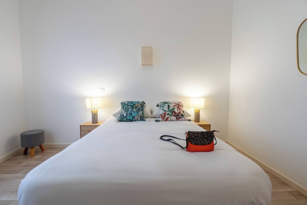 Un lit bien fait, avec deux oreillers, un petit tabouret et un sac à main posé sur le lit. Il y a deux lampes de chevet et une petite table avec une lampe.