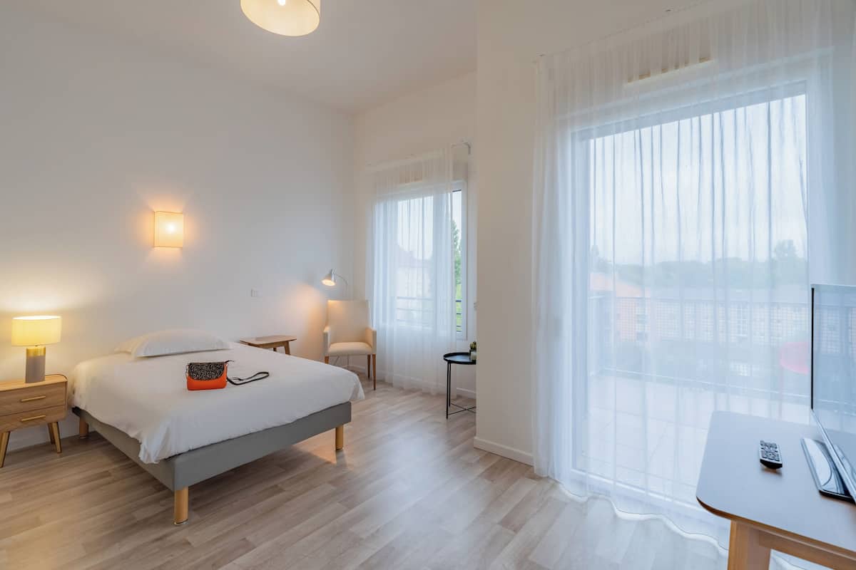 A hotel room with a bed, bedside table, lamp, chair, and a desk with a computer and remote control.