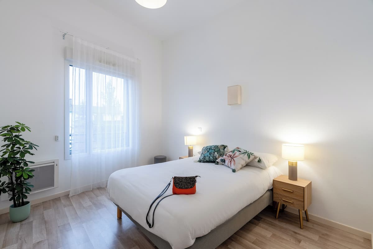 A bedroom with a bed, bedside lamps, a potted plant, and a window with sheer curtains.