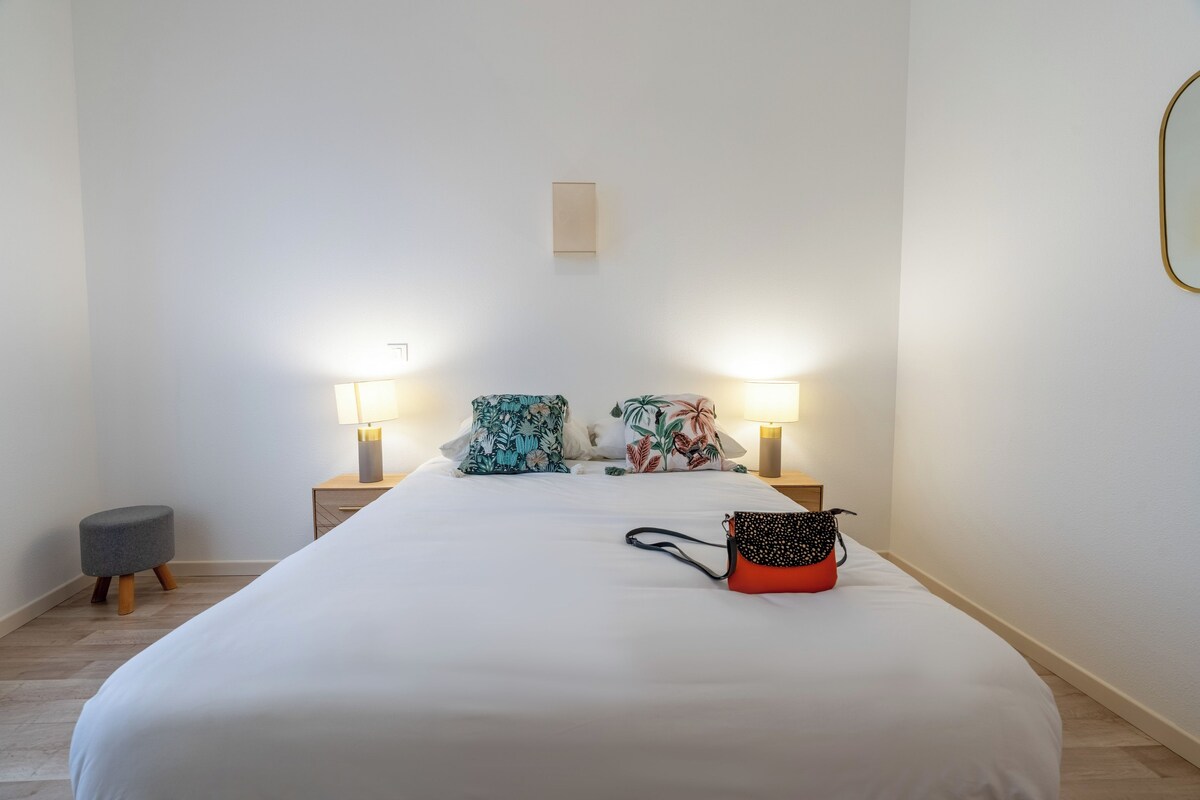 A neatly made bed with two pillows, a small stool, and a handbag on the bed. There are two bedside lamps and a small table with a lamp.