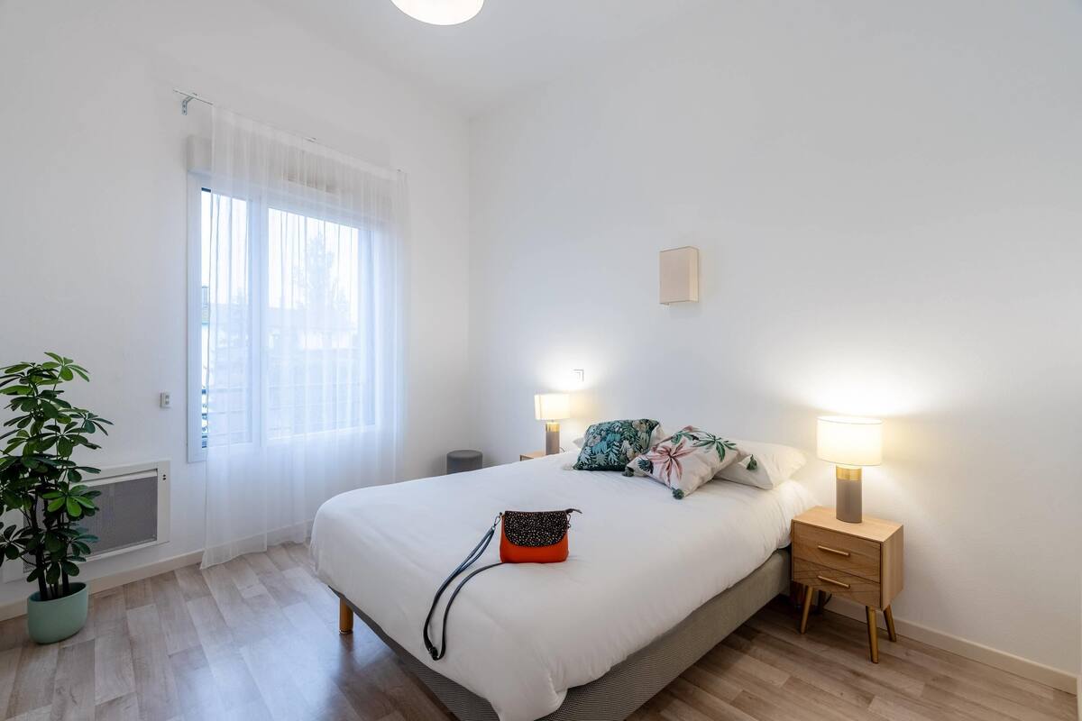 A bedroom with a bed, bedside tables, a lamp, a potted plant, and a window with sheer curtains.