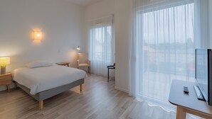 A hotel room with a bed, bedside table, lamp, chair, TV, and large windows. - Quality Aparthotel Mulhouse (Mulhouse)