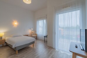 A hotel room with a bed, bedside table, lamp, chair, TV, and large windows. - Quality Aparthotel Mulhouse (Mulhouse)