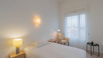 Een hotelkamer met een bed, nachtkastje, lamp, stoel en een klein tafeltje met een plant.