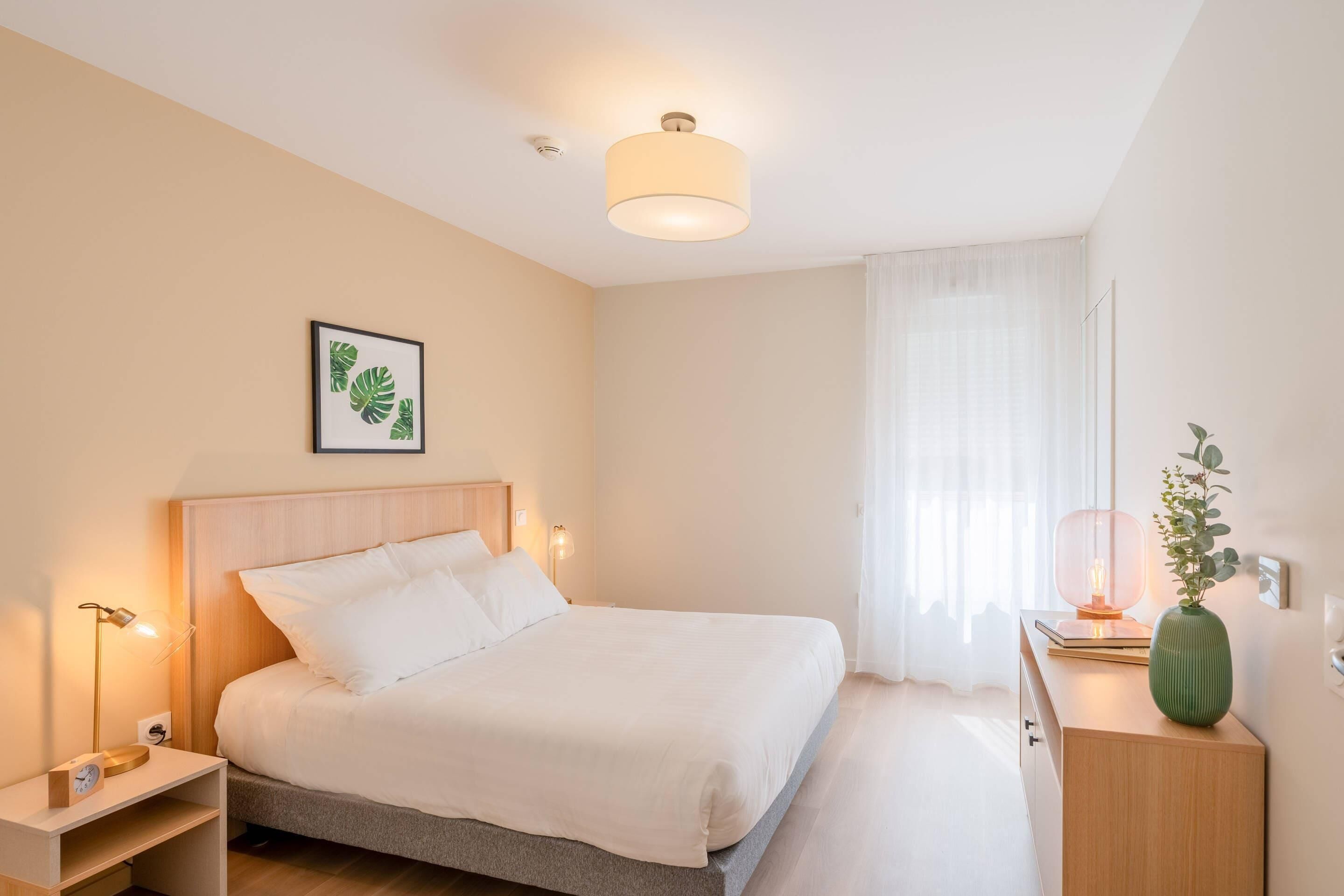 A neatly arranged bedroom with a bed, bedside lamps, a bedside table, a nightstand, a bedside clock, a framed picture on the wall, a window with sheer curtains, and a small green vase on a nightstand.