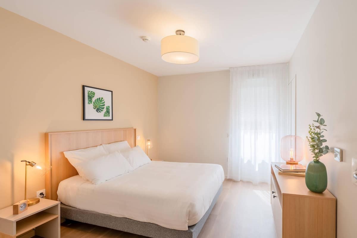 A neatly arranged bedroom with a bed, bedside lamps, a bedside table, a nightstand, a bedside clock, a framed picture on the wall, a window with sheer curtains, and a small green vase on a nightstand.
