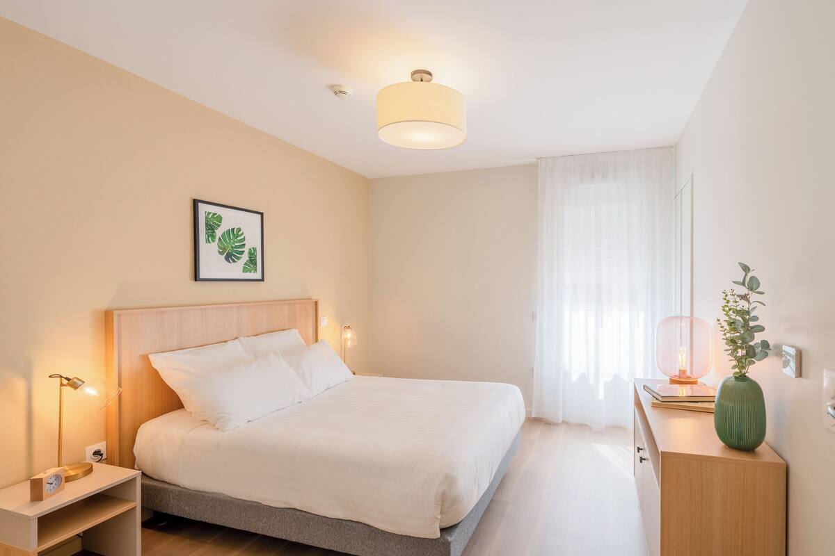 A neatly arranged bedroom with a bed, bedside lamps, a bedside table, a framed picture, and a window with curtains.