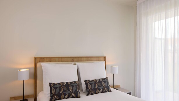 Una cama bien hecha con sábanas blancas y dos almohadones decorativos, flanqueada por mesitas de noche con lámparas, en una habitación con cabecera de madera y cortinas translúcidas.