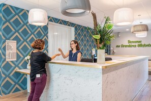 A reception area with a marble counter, a potted plant, and a sign indicating the location of various services. - Quality Aparthotel Villefontaine (Villefontaine)