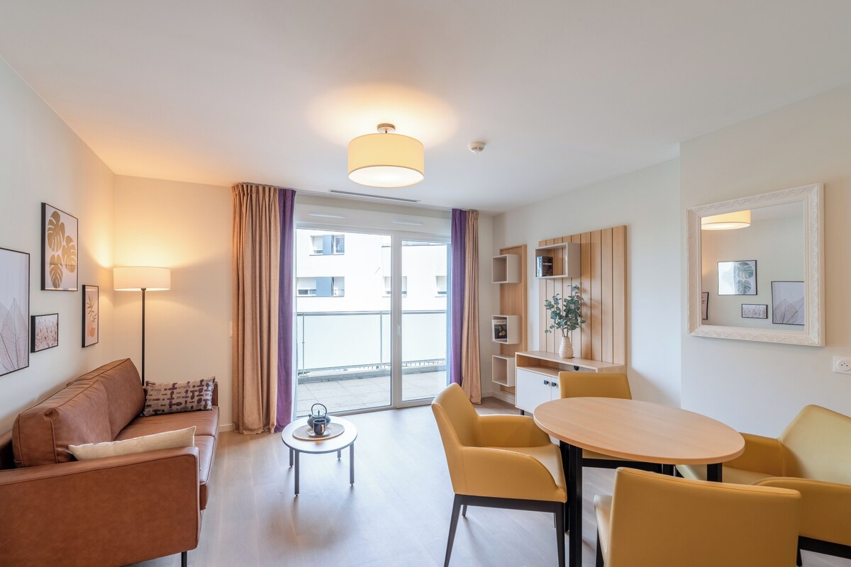 A modern hotel room with a sofa, a small table, a dining table with chairs, a lamp, a mirror, and a sliding door to the outside.
