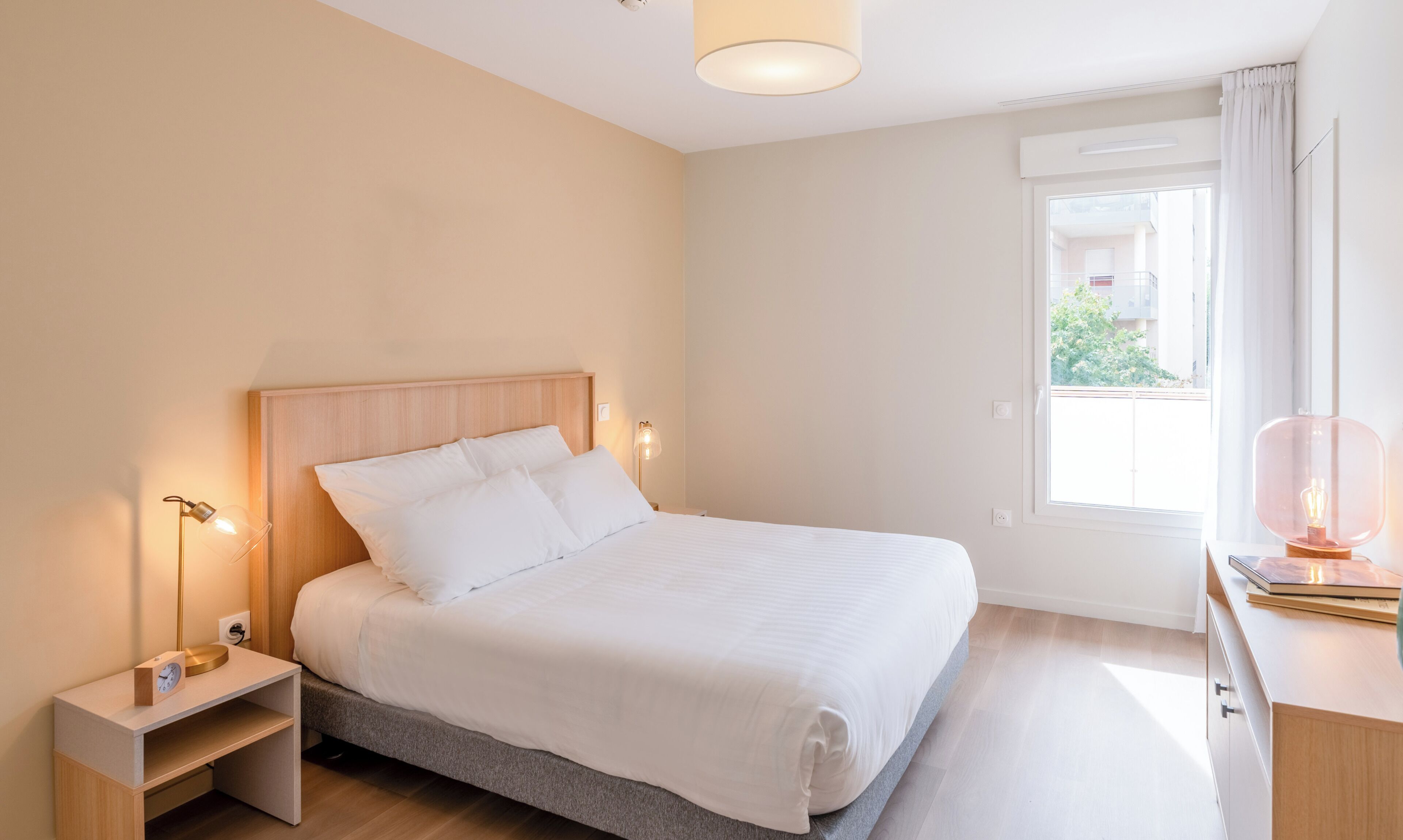 Une chambre à coucher comprenant un lit, des lampes de chevet, une table de chevet, une fenêtre avec des rideaux et un bureau avec une lampe.