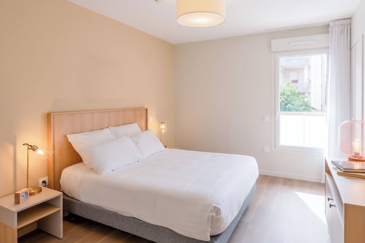 A bedroom with a bed, bedside lamps, a nightstand, a window with curtains, and a desk with a lamp.
