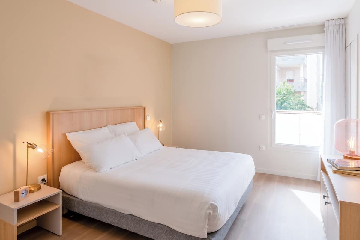 Une chambre à coucher comprenant un lit, des lampes de chevet, une table de chevet, une fenêtre avec des rideaux et un bureau avec une lampe.