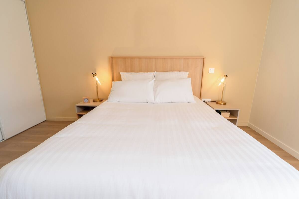 Un lit bien fait, avec du linge de lit blanc, deux tables de chevet avec des lampes et une tête de lit en bois.
