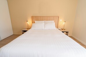 A neatly made bed with white linens, two bedside tables with lamps, and a wooden headboard. - Quality Aparthotel Villefontaine (Villefontaine)