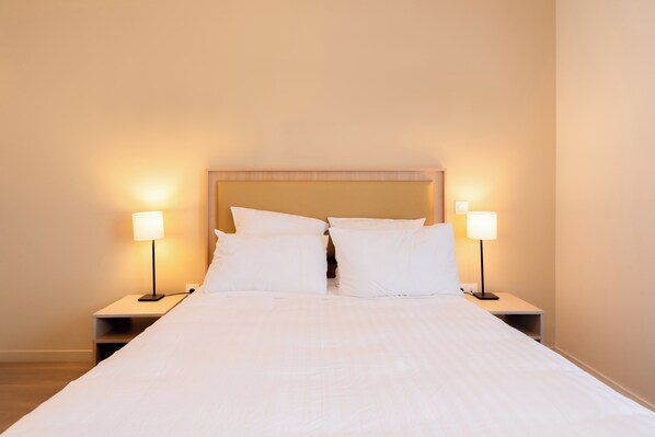 A neatly made bed with white linens, two bedside lamps, and a wooden headboard. - Quality Aparthotel Villefontaine (Villefontaine)