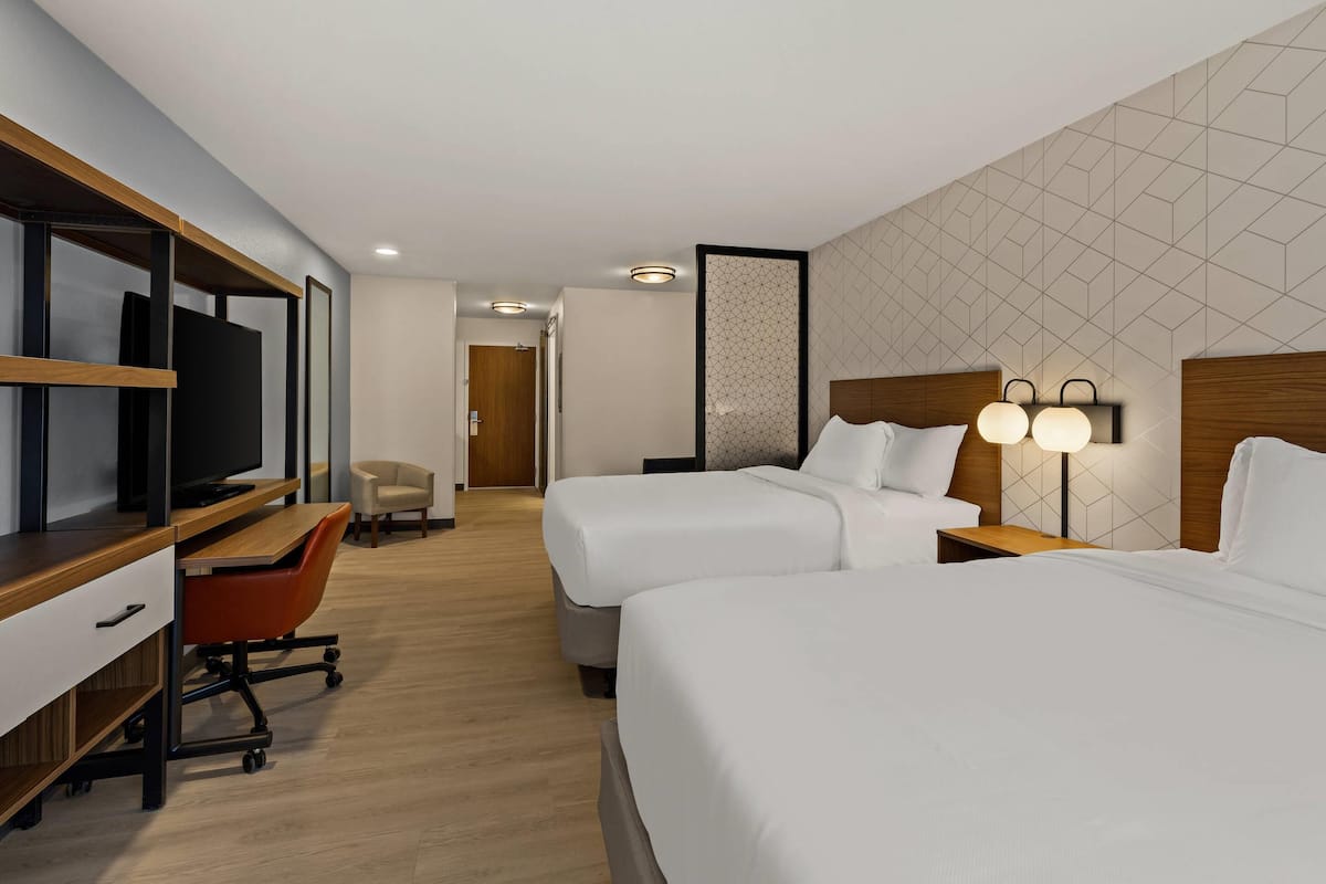 Ein Hotelzimmer mit zwei Betten, einem Schreibtisch mit Stuhl, einem Fernseher und einer Wandlampe.