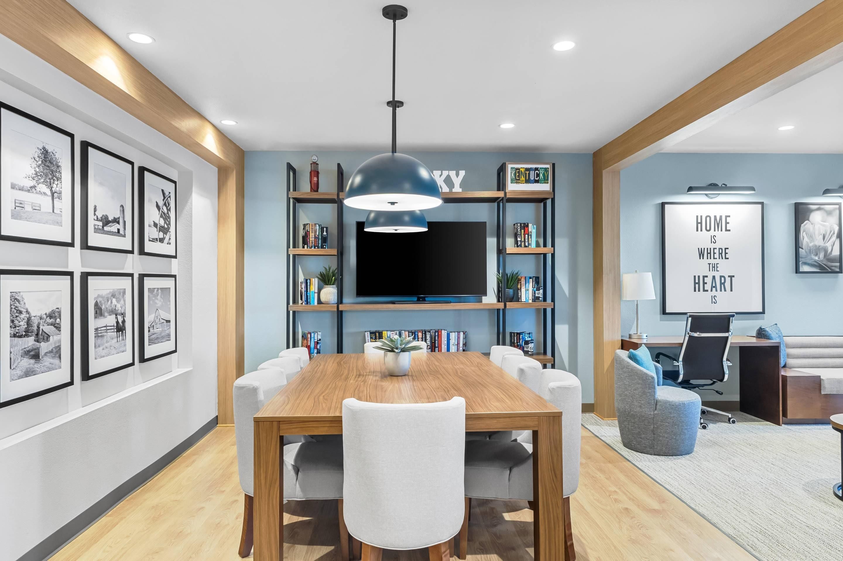 A modern dining area with a wooden table and chairs, a large flat-screen TV, and a bookshelf with various items.