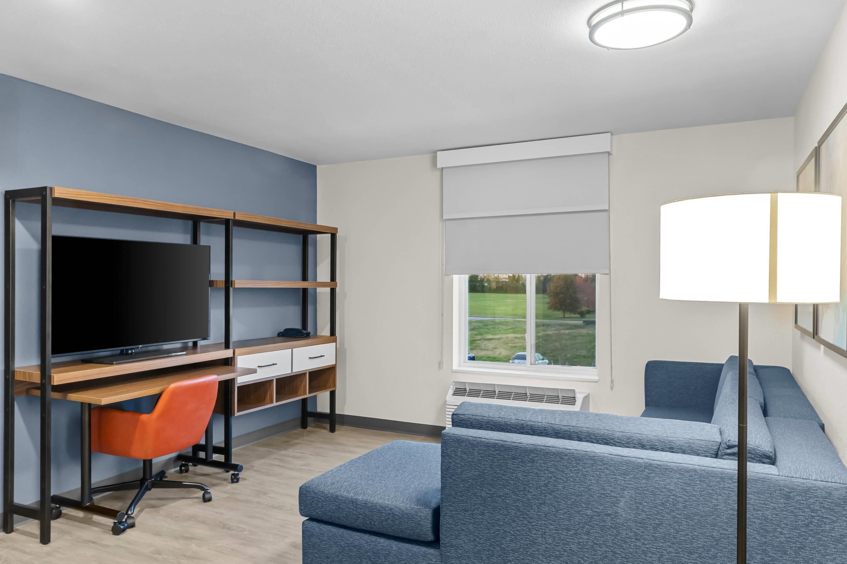 A modern hotel room with a flat-screen TV, a desk with a chair, a sofa, a standing lamp, and a window with a view of the outdoors.