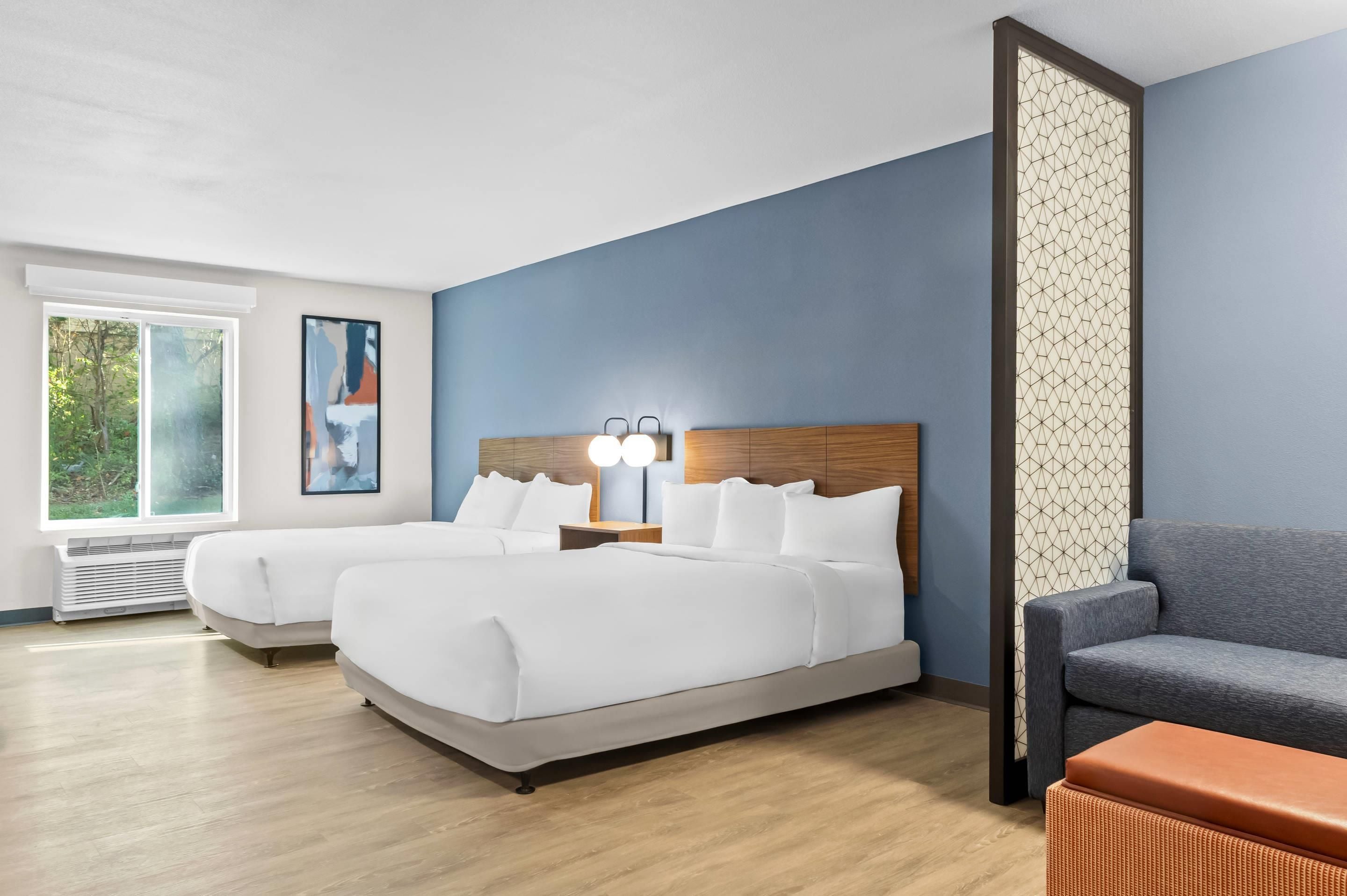 Ein Hotelzimmer mit zwei Betten, einem grauen Sofa, einem hölzernen Kopfteil, einem Nachttisch mit Lampe und einem Fenster mit Blick auf Grünflächen.