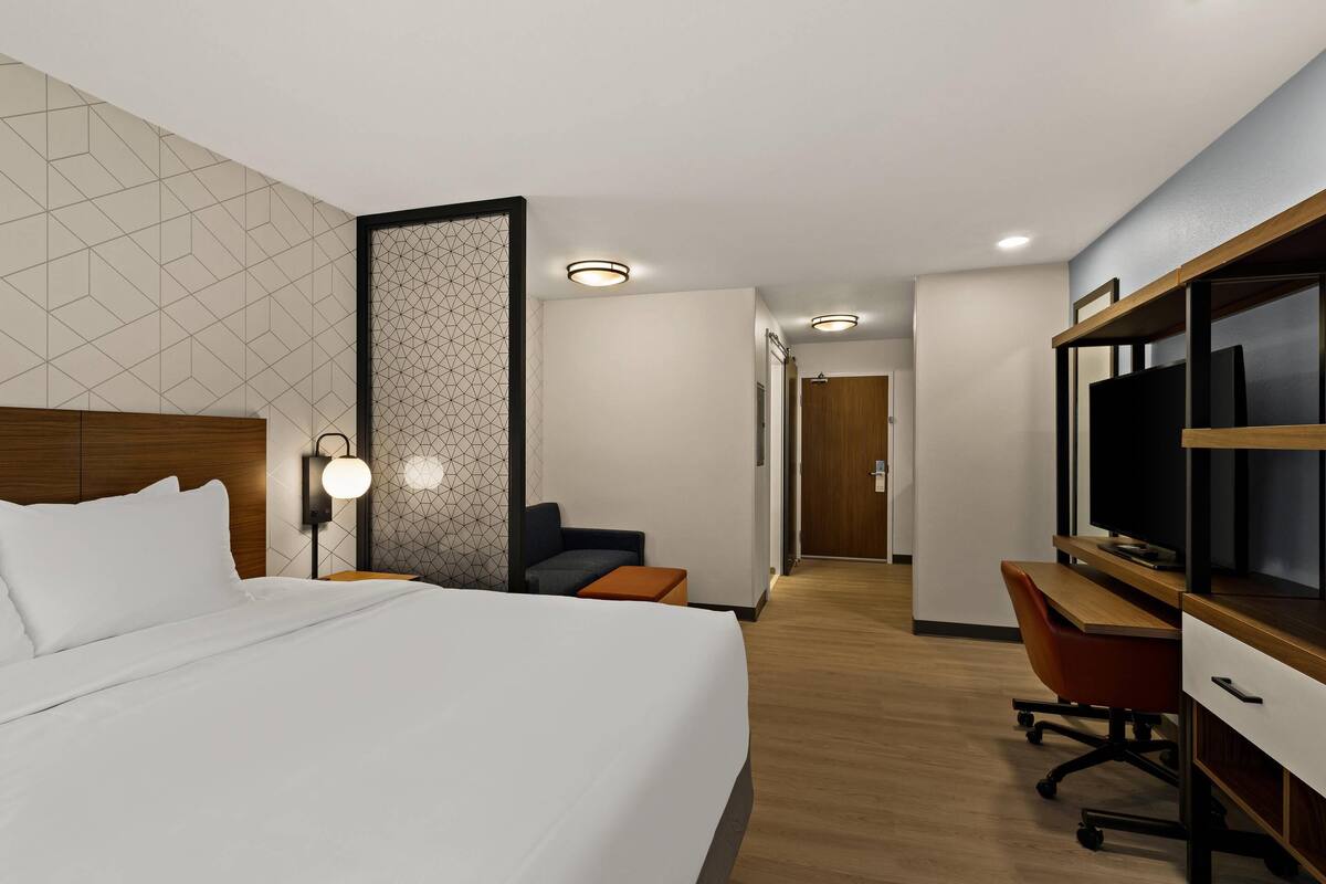 Ein modernes Hotelzimmer mit einem großen Bett, einem Schreibtisch mit Computer, einem Fernseher und einer Sitzecke mit Sessel und kleinem Tisch.