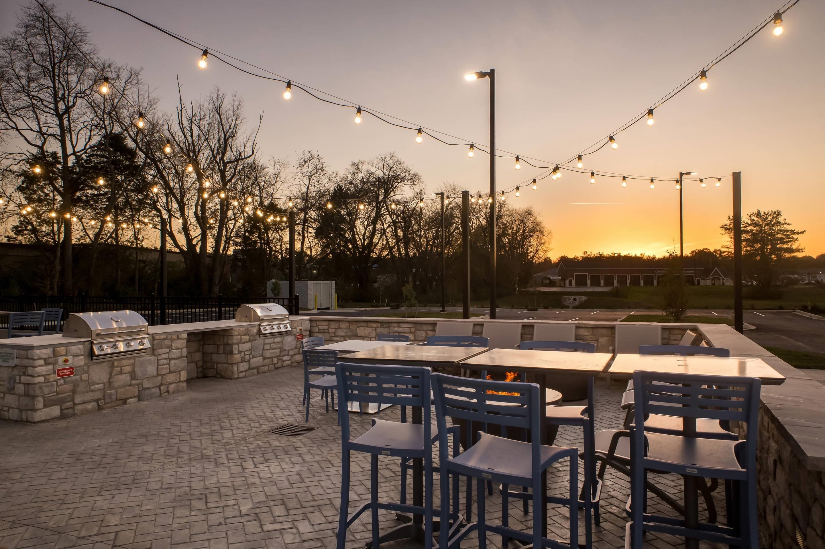 Un patio al aire libre con asientos de piedra y una parrilla, luces colgantes y un atardecer de fondo.