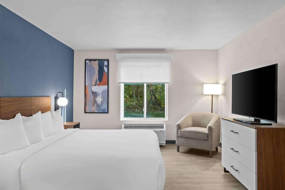 Ein Hotelzimmer mit einem großen Bett, einem Fernseher, einem Sessel und einem Fenster mit Blick auf Grünflächen.
