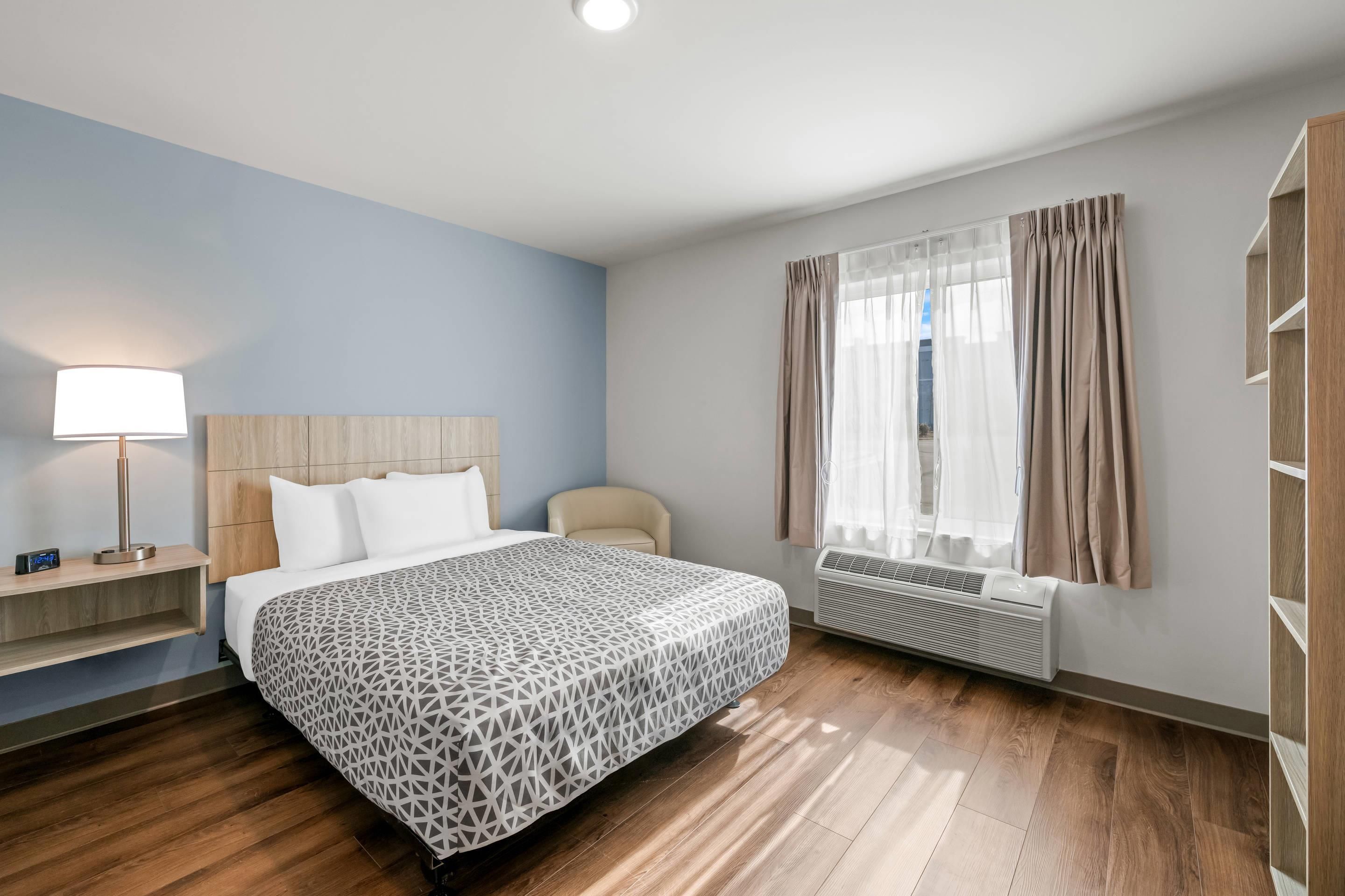 Hotelkamer met een bed, nachtkastje, lamp, stoel, raam met gordijnen en een ingebouwde airconditioning.