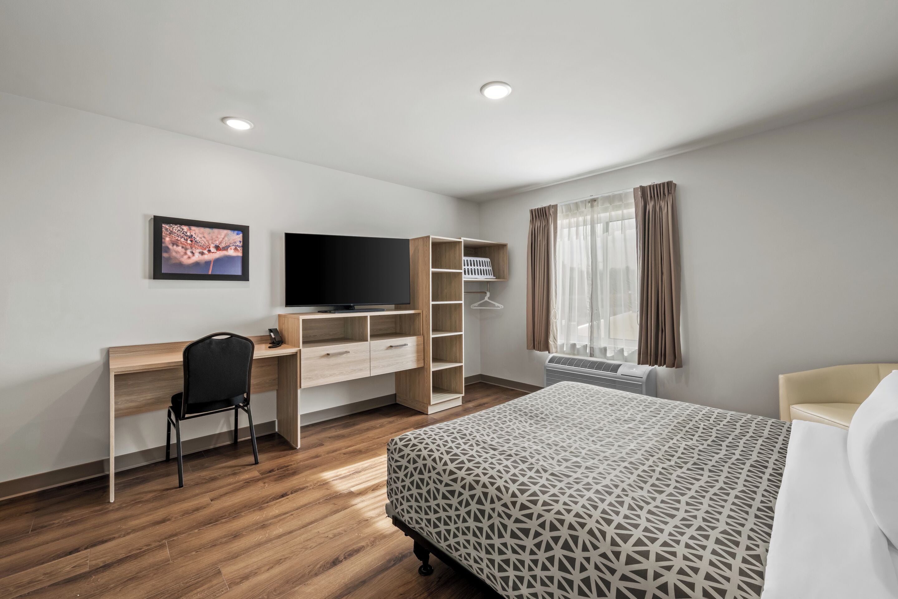 A modern hotel room with a wooden desk, a black chair, a bed with a geometric patterned bedspread, a television, and a wardrobe.