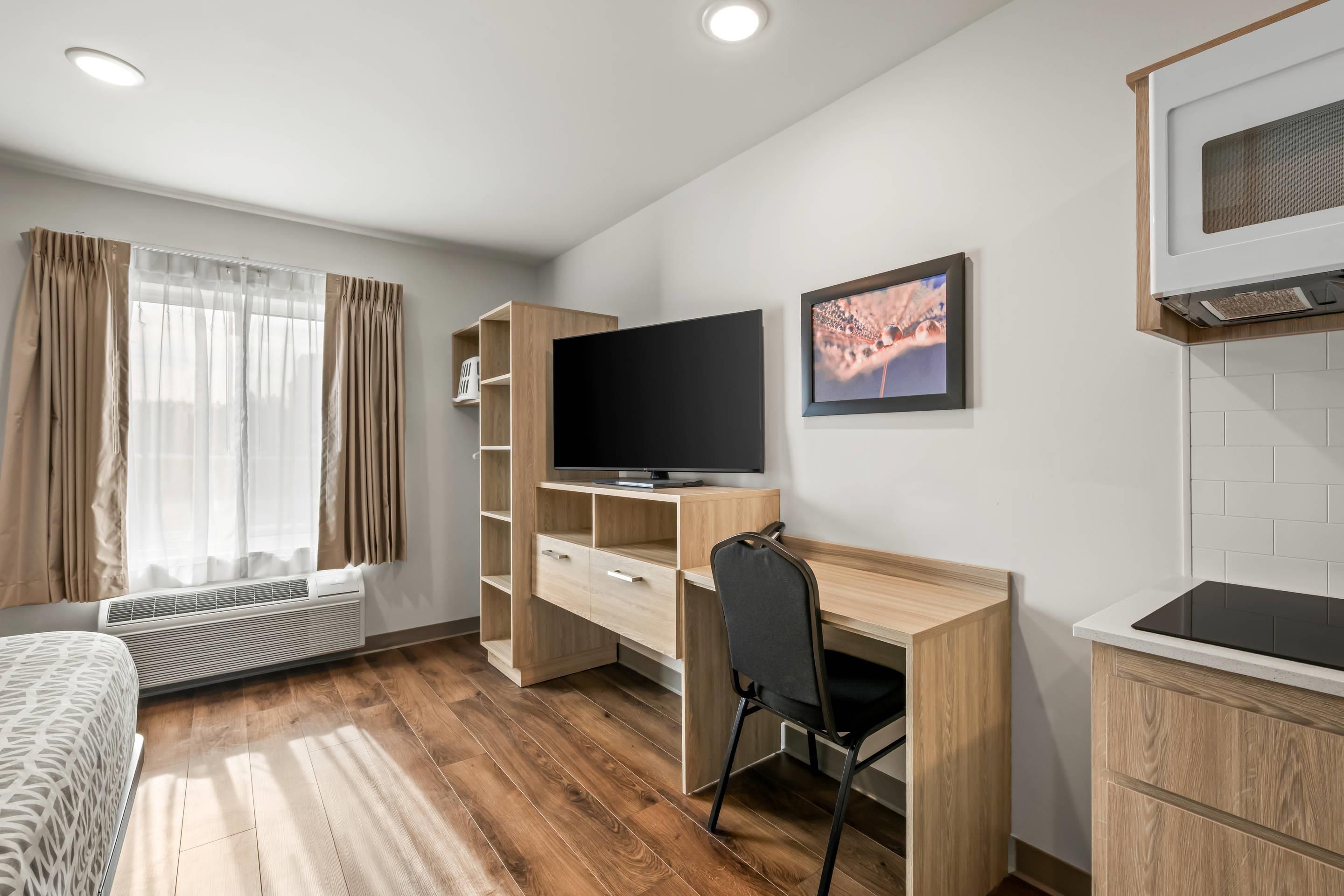 Moderni hotellihuone, jossa on puinen työpöytä, taulutelevisio, sänky ja pieni keittiönurkkaus.