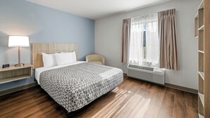 Una habitación de hotel moderna con una cama grande, una mesita de noche con lámpara, una silla, una ventana con cortinas y un equipo de aire acondicionado integrado.