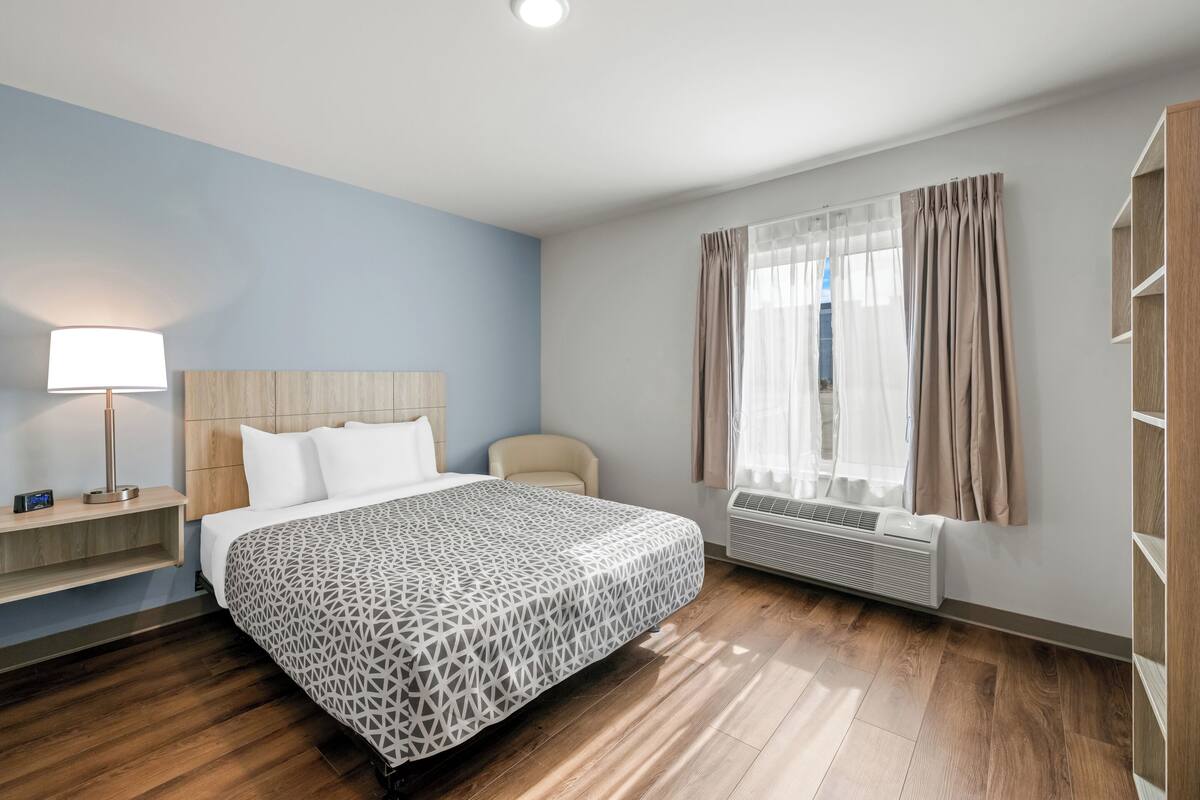 Ein modernes Hotelzimmer mit einem großen Bett, einem Nachttisch mit Lampe, einem Sessel, einem Fenster mit Vorhängen und einer eingebauten Klimaanlage.
