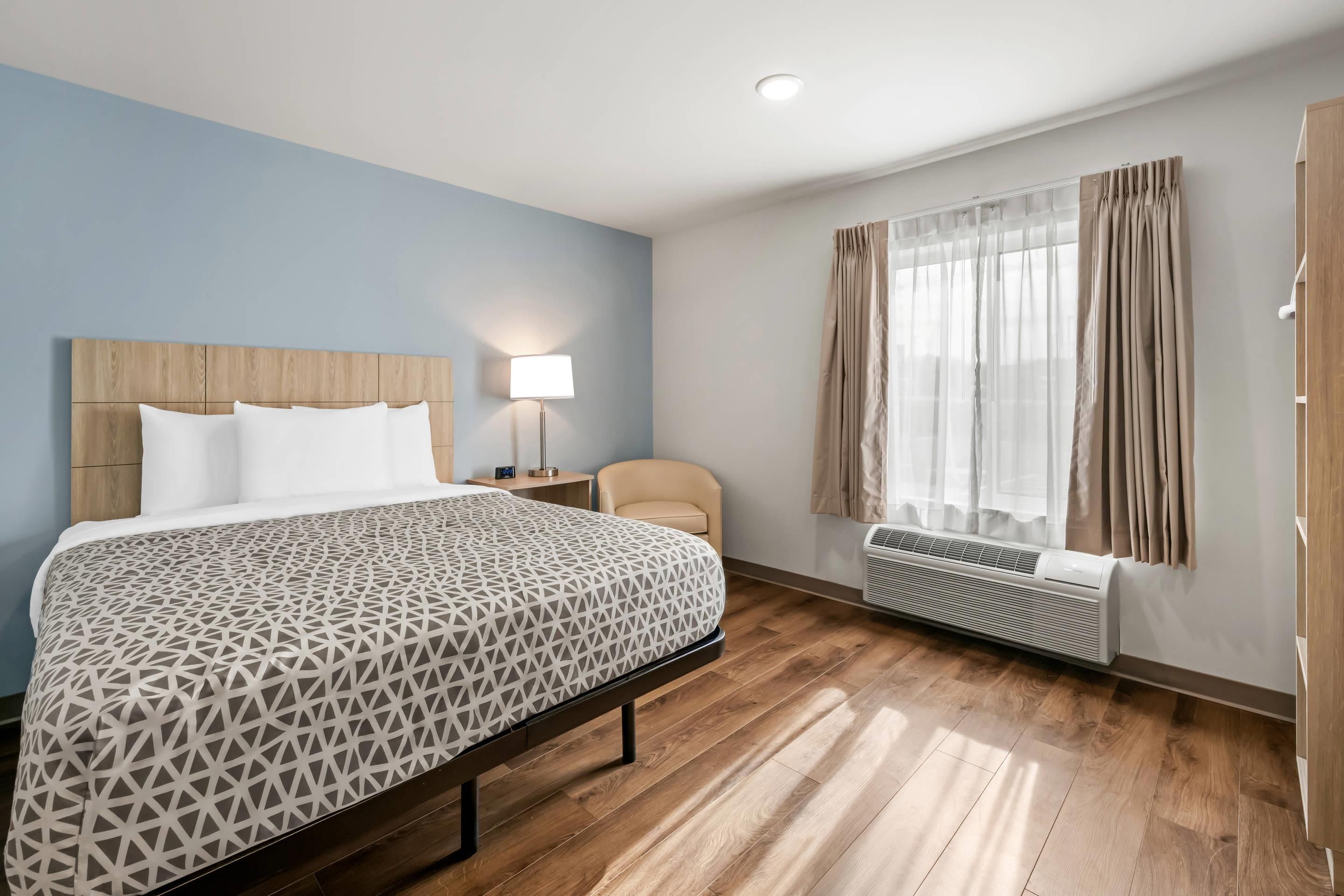 Ein Hotelzimmer mit Bett, Sessel, Nachttisch, Lampe, einem Fenster mit Vorhängen und einer Klimaanlage.