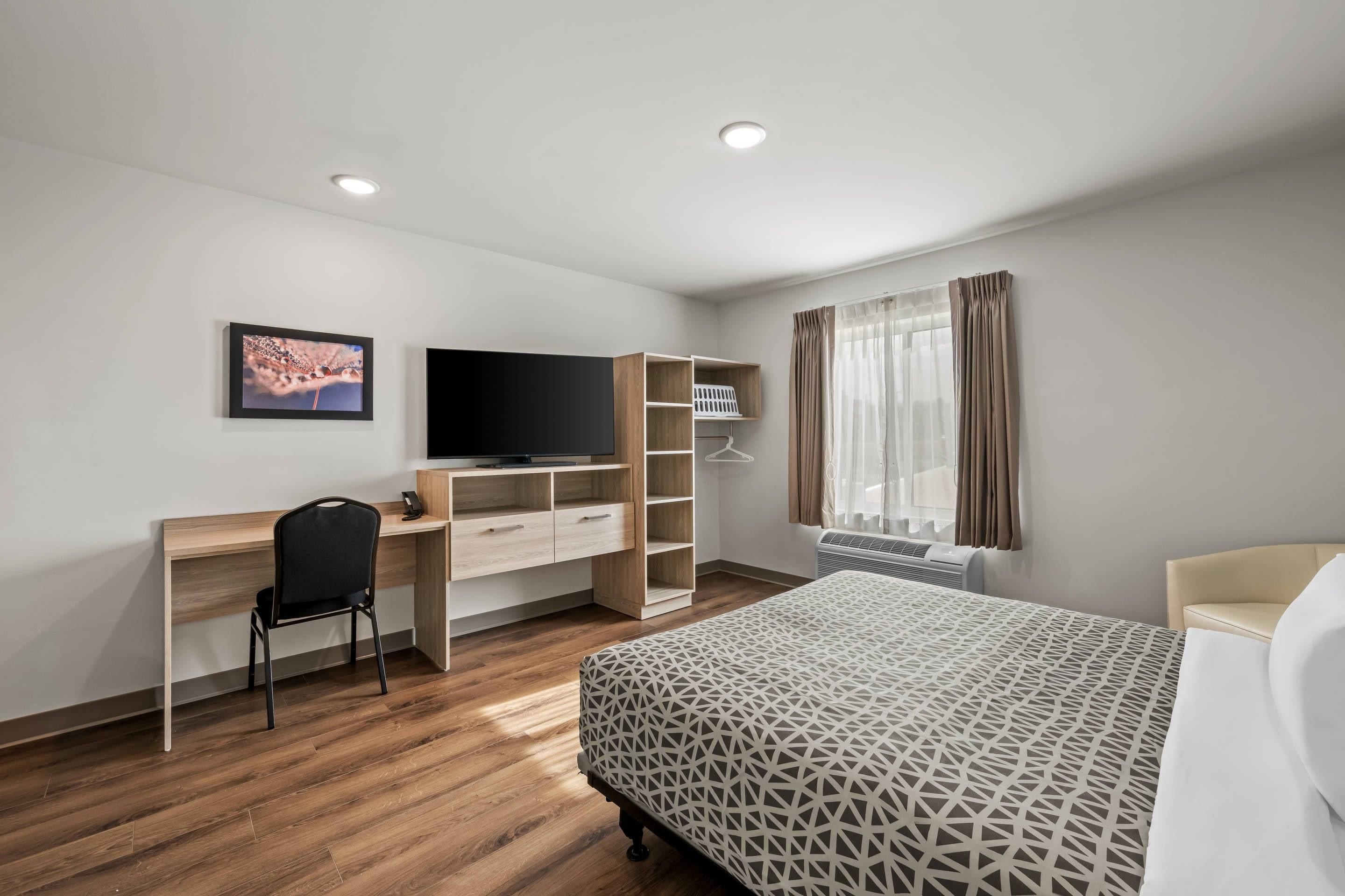 Une chambre d’hôtel moderne avec un bureau en bois, une chaise noire, un lit recouvert d’une courtepointe à motifs géométriques, une télévision et une fenêtre avec des rideaux.