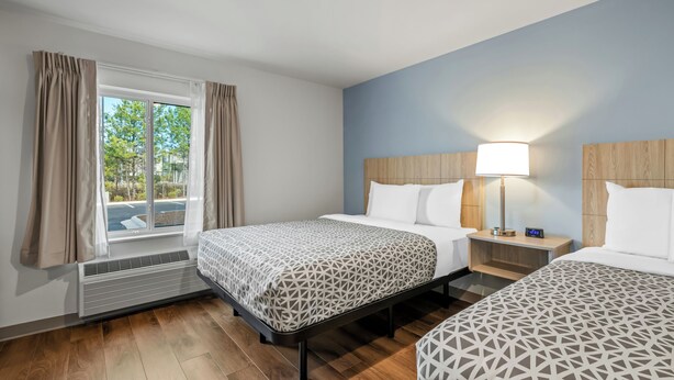 A hotel room with two beds, a window with curtains, a bedside table with a lamp, and a heater. - WoodSpring Suites Charlotte Northlake (Charlotte)