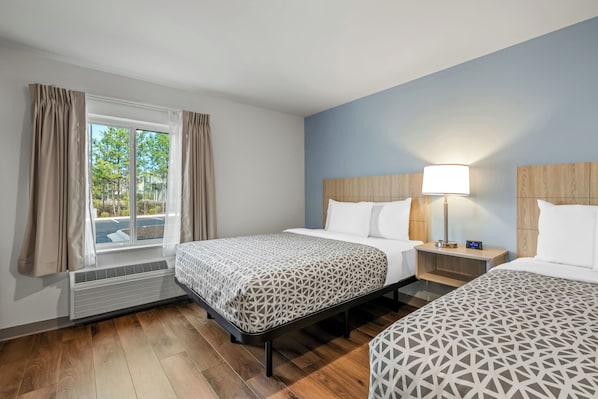 A hotel room with two beds, a window with curtains, a bedside table with a lamp, and a heater. - WoodSpring Suites Charlotte Northlake (Charlotte)