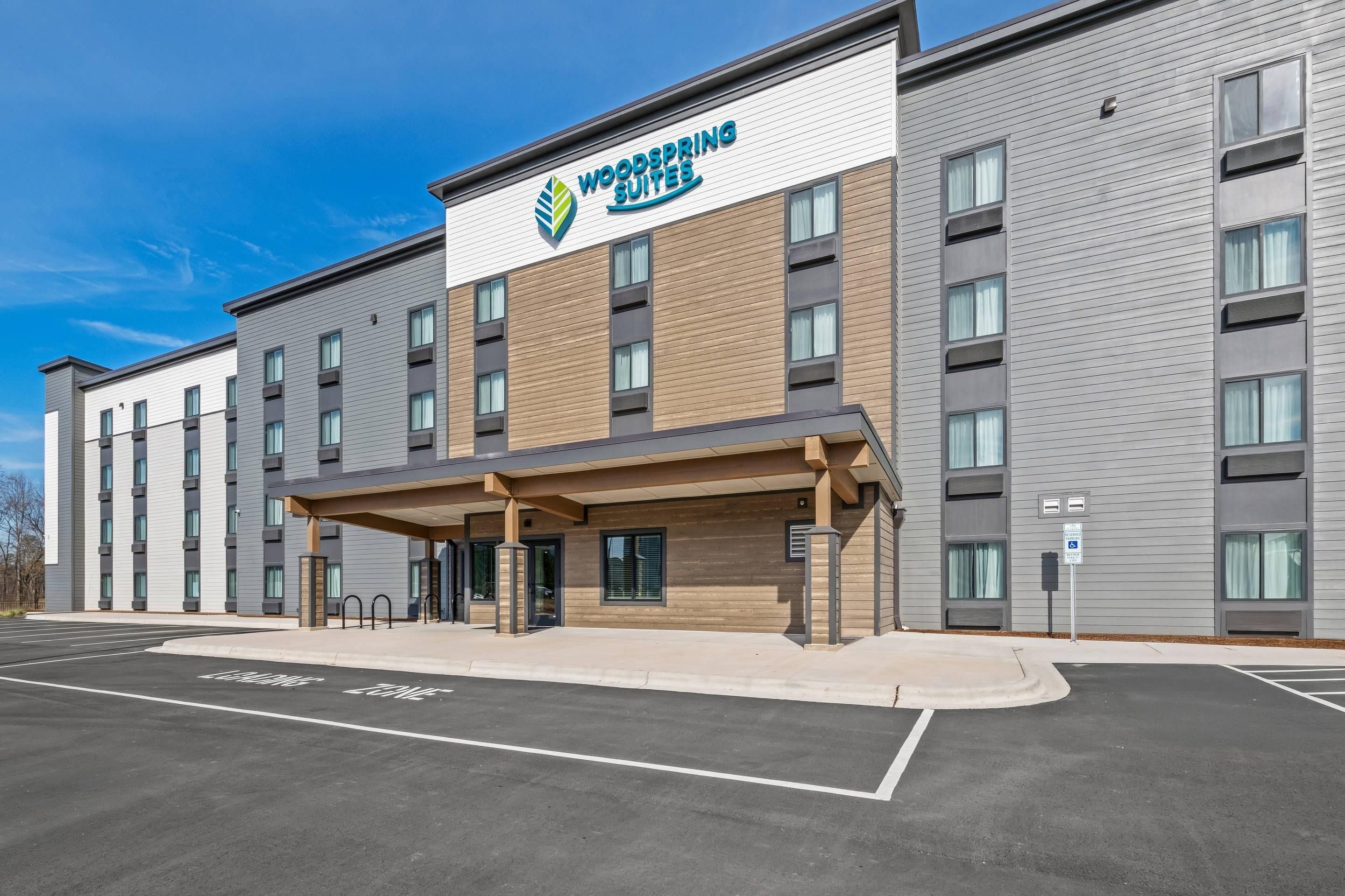 Ein modernes Hotelgebäude mit der Aufschrift 'Woodspring Suites'. Es verfügt über einen überdachten Eingang, zahlreiche Fenster und einen Parkplatz mit markierten Stellplätzen.
