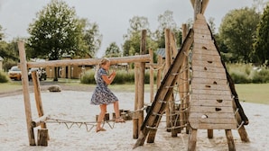 A wooden play structure with a rope bridge and a teepee-like building on a sandy beach. - Charming Tiny House Getaway With Terrace (Balkbrug)
