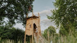 A wooden treehouse with a ladder leading up to it, surrounded by greenery. - Charming Tiny House Getaway With Terrace (Balkbrug)