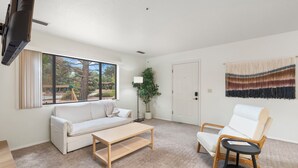 Un salon spacieux avec un canapé blanc, une table basse en bois et un fauteuil blanc. Une grande fenêtre offre une vue sur l’extérieur.