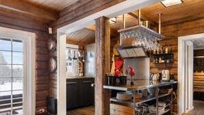 L’intérieur d’une cabane en rondins comprend un espace cuisine avec un bar en acier inoxydable, un plancher en bois et une vue à travers une porte vitrée.