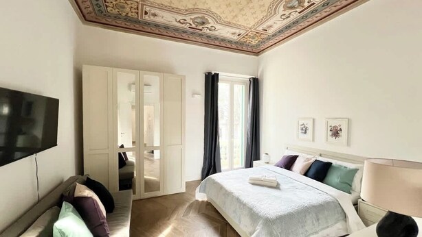 Quarto de hotel com cama, televisão, guarda-roupa e teto com detalhes elaborados.