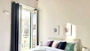 Quarto com cama, mesa de cabeceira, luminária e janela com cortinas.