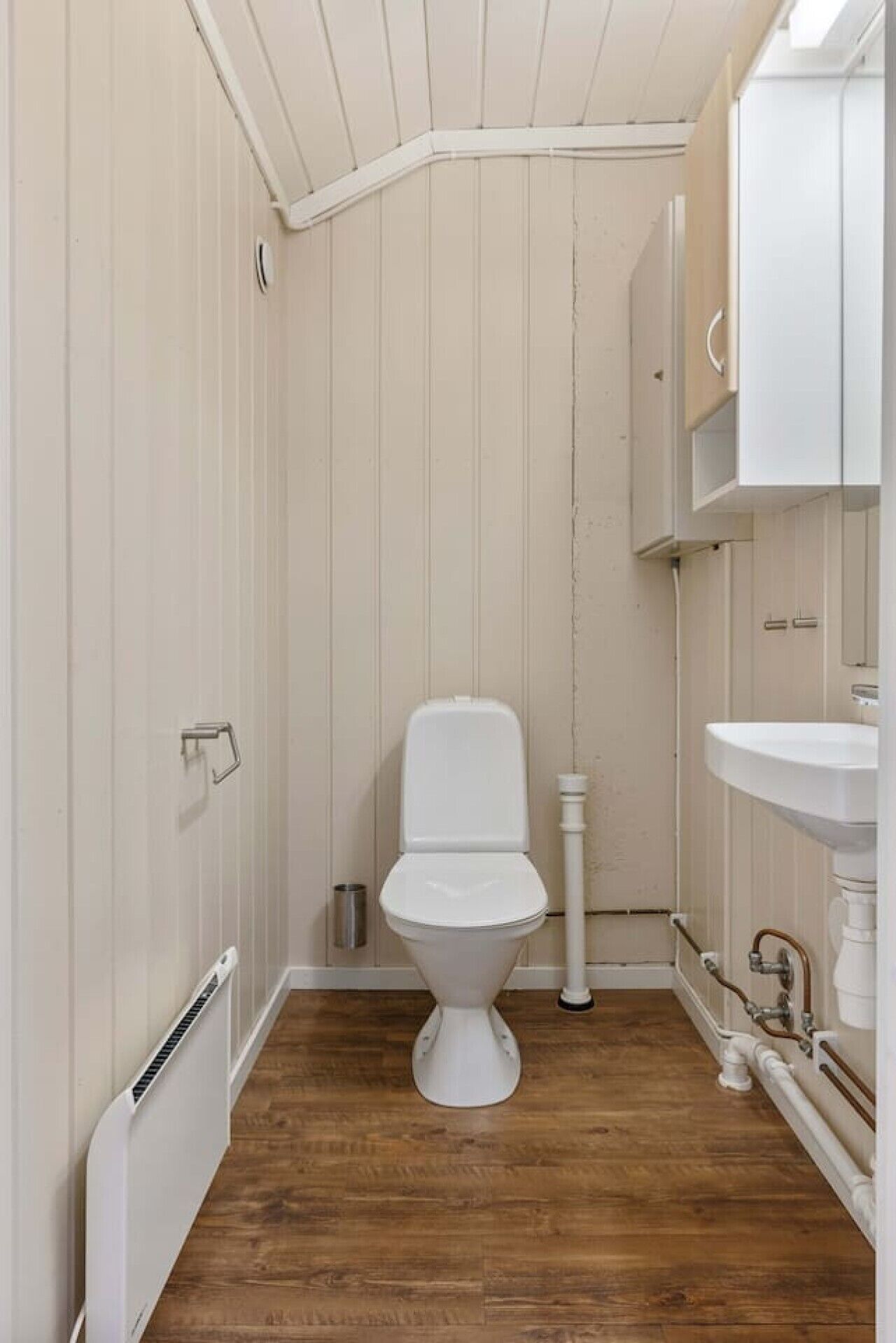 Et lille, smalt badeværelse med et hvidt toilet, trægulv og en hvid vask.