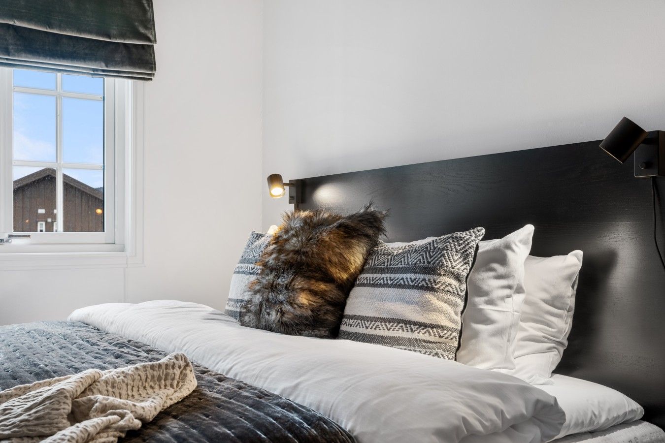A bedroom with a bed, pillows, a blanket, a bedside lamp, and a window with a view of buildings.