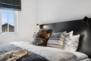 A bedroom with a bed, pillows, a blanket, a bedside lamp, and a window with a view of buildings.