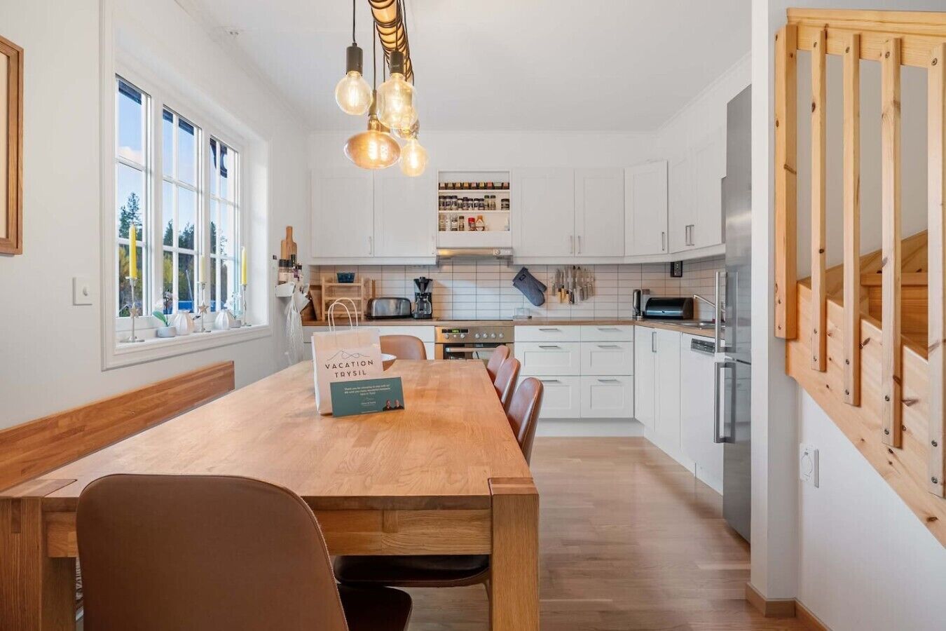 A modern kitchen with white cabinets, a wooden dining table, and a hanging light fixture.