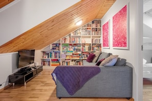 A loft-style room with a sloped ceiling, a built-in bookshelf, a grey sofa, and a television. - Trysil Ski-in out Apt, Private Sauna, Sleeps 9 (Trysil)