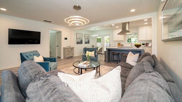 Una sala de estar moderna con un sofá, una mesa de centro y un televisor. En el fondo, hay una zona de cocina con una barra y una mesa de comedor.